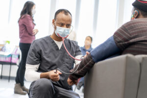 A volunteer wearing gray scrubs and a face mask provides a blood pressure check for a guest at the Augsburg Central Health Commons.